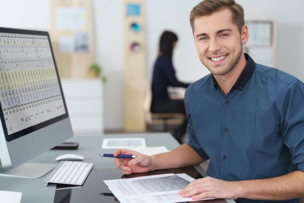 Homme souriant en chemise bleue, assis à un bureau avec un écran d'ordinateur affichant un tableau de données, tenant un stylo et des documents.
